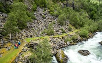 To vandrere i farverige jakker på den smalle Kongevegen-sti ved en rivende turkis flod i en klippefyldt norsk kløft med grøn vegetation.