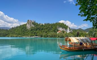 Traditionella träbåtar på det turkosa Bledsjön med Bled slott på en skogbeklädd klippa och ett vitt kyrktorn synligt.