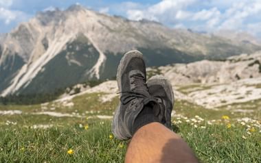 Grå vandrestøvler på krydsede ben hvilende i alpin eng med gule vilde blomster, dramatiske Dolomitternes toppe i baggrunden.