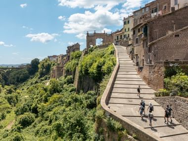 Gruppe vandrere på buet stibro til historiske Orvieto med gamle bygninger og grøn dal under blå himmel.