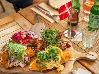 Träbräda med danska smörgåsar toppade med fisk, grönsaker och färska örter. En liten dansk flagga står i mitten.