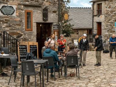 Gruppe Camino de Santiago pilgrimme med rygsække hviler ved café borde på historisk stentorv med traditionelle bygninger.