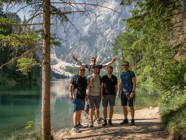 Fem vandrere poserer ved en turkis bjergsø i Dolomitterne, omgivet af tæt skov og dramatiske klippetoppe.