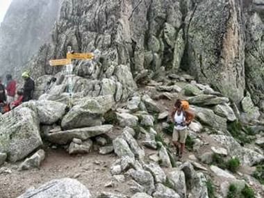 Vandrer med rygsæk på stenet terræn på Mont Blanc-ruten. Store granitblokke og vejskilt synlige i tågede bjergforhold.