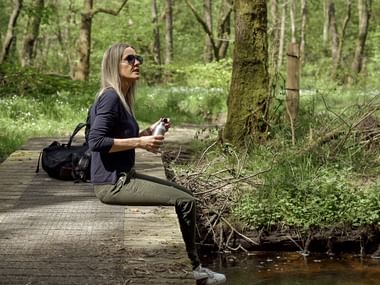 Kvinna sitter på träspång över bäck i dansk skog, bär solglasögon och håller telefon, omgiven av frodig grön vegetation.