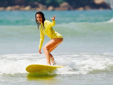 Kvinde i lysegul våtdragt surfer på gult surfbræt i turkise farvande ud for Sri Lanka og laver fredstegn med hånden.
