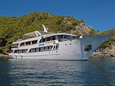 Vit motoryacht MY Mystic ankrad i lugna blå vatten längs den kroatiska kusten med skogklädda klippor i bakgrunden.