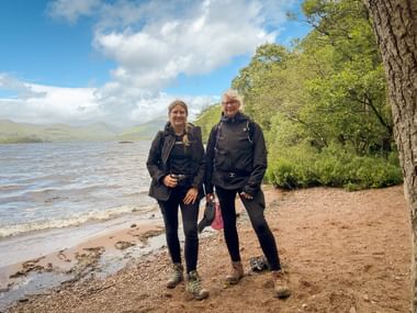 To vandrere i mørkt udendørstøj står på en sandstrand ved en sø på West Highland Way, med bakker og træer i baggrunden.