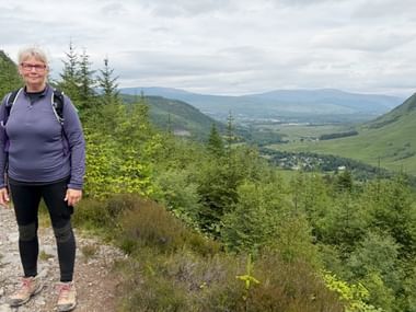 Kvinde med rygsæk og lilla fleece på grusvej med udsigt over grøn dal med bjerge i de skotske højlande.