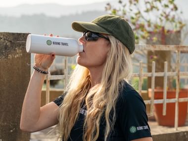 Kvinde med blondt hår, kasket og solbriller drikker fra hvid vandflaske på den portugisiske Camino med stenmure i baggrunden.