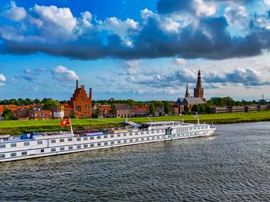 Hvidt MS Rigoletto flodkrydstogtskib sejlende på stille vand foran historisk hollandsk by med kirketårn og traditionelle bygninger under skyet himmel.