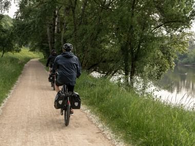 Cyklist med cykeltasker kører på grusvej langs Loire, omgivet af frodige grønne træer og græs på en fredelig landevej.