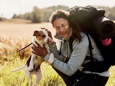 Smilende kvinde med rygsæk knæler i gyldent felt, omfavner lille brun-hvid hund i snor. Bakker og træer i baggrunden.