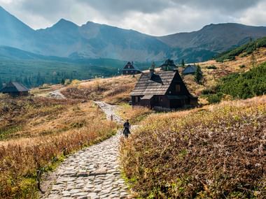 Stenstien fører til traditionelle træhytter ved Hala Ornak i Tatra-bjergene, med vandrere på stien gennem efterårsenge.
