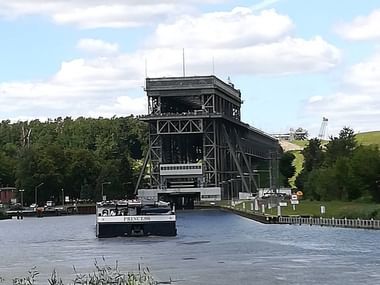 Hvidt passagerskib sejler gennem hævet stål løftebro på rolig vandvej, omgivet af grønne træer og skyet himmel.