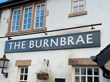 Hvid bygning med stendetaljer og traditionelle vinduer med 'The Burnbrae'-skilt på facaden, med hængende kurv og gadelampe.