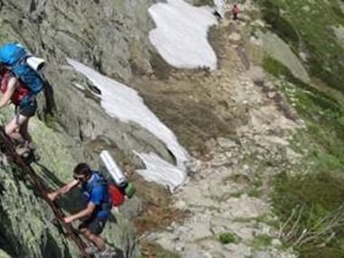 Vandrere med rygsække klatrer på metalstiger på en stejl klippefyldt bjergsti med snepletter. Stien slynger sig op ad bjerget.