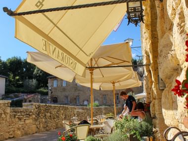 Udendørs restaurantterrasse med cremefarvede parasoller og stenvægge. En person sidder ved et bord i skyggen, med røde blomster og historiske bygninger synlige.