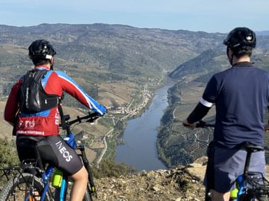 To cyklister med hjelme og cykler holder pause på en bakke med udsigt over den snoede Douro-dal med terrasserede vingårde.