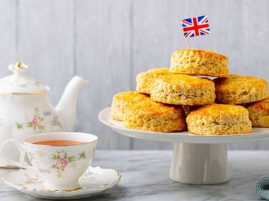Stapel gyllene scones på vit tårtfat med Union Jack-flagga på toppen, tekopp med blommönster och matchande tekanna på marmoryta.
