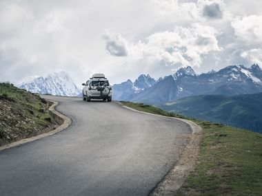 SUV med tagbagagebærer på snoet bjergvej. Snedækkede tinder synlige i baggrunden under overskyet himmel med grønne skråninger.