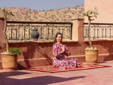 Kvinde i lyserød mønstret kjole sidder i meditationsstilling på farverigt tæppe på terrasse med Atlas-udsigt, lerpotter og jerngelænder.