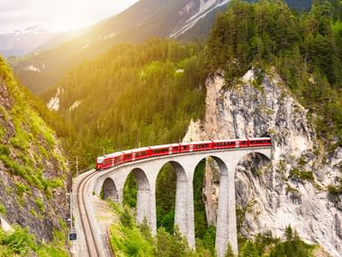 Rødt passagertog krydser en buet stenviadukt med flere buer gennem en schweizisk bjergdal med tætte skove og klippevægge.
