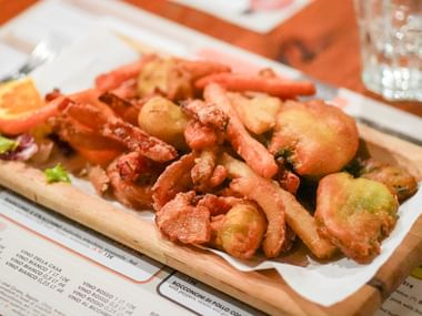 Træbræt med friterede skaldyr inklusiv rejer, blæksprutter og grøntsager, serveret på hvidt papir over en menu på orange dug.