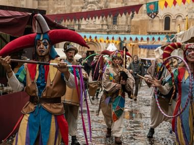 Middelalderlige kunstnere i farverige narreskostymer med bjælder og bånd, der danser på en historisk stentorv pyntet med festgirlander.