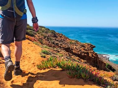 Hiker with backpack walking on orange sandy cliff path along Portuguese coast with blue ocean and rocky cliffs covered in green vegetation.