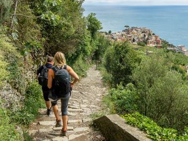 To vandrere med rygsække på stenet sti med udsigt over Cinque Terre landsby og Middelhavet, omgivet af frodig vegetation.