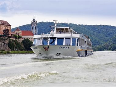 Hvidt flodkrydstogtskib MS SE-MANON sejlende på Donau med historisk by og barok kirketårn på skråning i baggrunden.