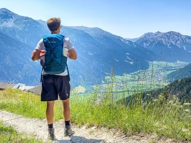 Vandrer med blå rygsæk på bjergsti med udsigt over Alpedal med grønne enge og snedækkede toppe ved Garmisch.