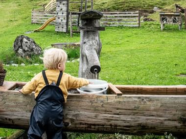 Blondt barn i gul trøje ved rustikt trævandtrug med udskåret fontæne i Salzkammergut. Grøn eng og legeplads i træ i baggrunden.