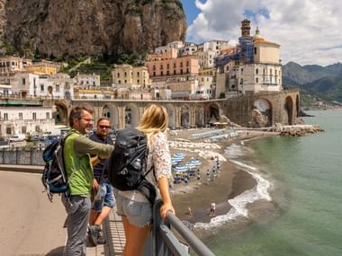 Three tourists with backpacks standing at viewpoint overlooking coastal town built into cliffs with beach below and turquoise sea.