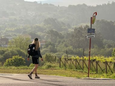 Kvindelig pilgrim med rygsæk går på vej forbi Camino-vejviser nær Rubiáns, med vingård og grønne bakker i baggrunden.