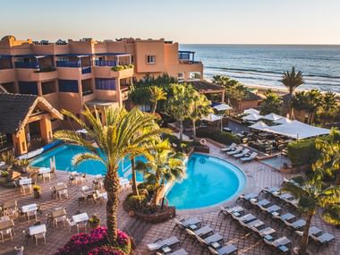Strandresort med pools i Agadir Luftfoto af Paradise Plage resort i Agadir med buede pools, palmer, solstole og strandplacering ved solnedgang.