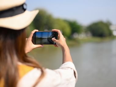 Kvinde i stråhat tager foto af sølandskab med smartphone. Grønne træer langs bredden under klar blå himmel.