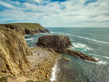 Dramatisk Cornwall-kustlinje med gyllene sandstensklippor, isolerad klippstack i turkost hav och vita skumvågor under molnig himmel.