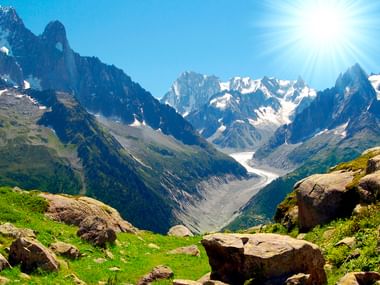 Två vandrare står på stenig terräng med utsikt över Mont Blanc massivet med snötäckta toppar, glaciärer och gröna alpängar.