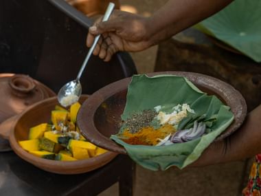 Hænder forbereder srilankansk curry med krydderier i en lerskål foret med bananblad, ved siden af mangostykker i en træskål.