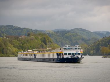 MS Prinzessin Katharina flodkrydstogtskib sejler på stille vand med skovklædte bakker i baggrunden under en overskyet himmel.