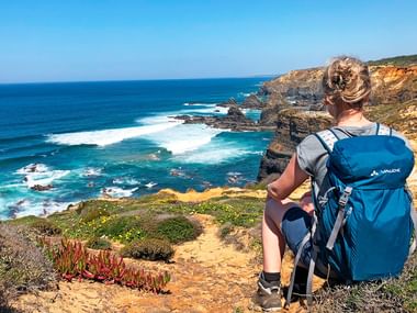 Hiker with blue backpack sitting on cliff overlooking Atlantic Ocean on Rota Vicentina trail. Dramatic coastline with waves and vegetation visible.