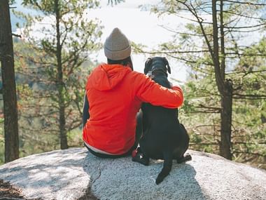 Person i rød jakke og hue sidder på stor klippe med sort hund, omgivet af skovtræer med bjerglandskab i baggrunden.