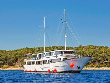 Hvid motoryacht MS Azimut med to master for anker i blåt vand nær den skovklædte kroatiske kyst under klar blå himmel.