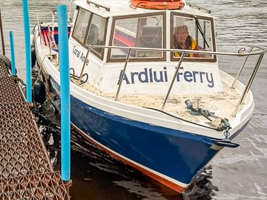 Lille færge ved navn Ardlui Ferry fortøjet ved pier med orange redningskranse på taget. Bjerge og sø synlige i baggrunden, Skotland.
