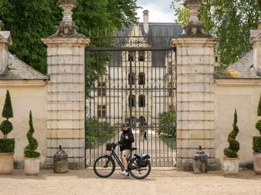 Cyklist med cykel vid smidesjärnsgrindarna till franskt slott med stenpelare, formklippta träd och klassisk arkitektur.