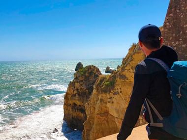 Hiker with blue backpack standing on golden cliffs overlooking turquoise ocean waves and rock formations along the Algarve coast.