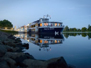 MS Princess krydstogtskib fortøjet ved kaj under aftenskumring, med varme lys fra vinduer og perfekt reflektion i roligt vand.
