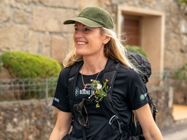 Smilende vandrer med grøn kasket og rygsæk iført en sort Bering Travel trøje, går gennem en portugisisk landsby med stenbygninger.